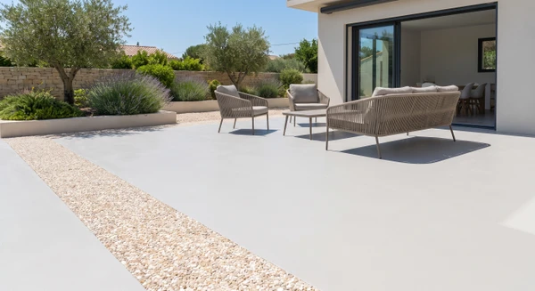 Terrasse extérieure résidentielle avec revêtement de sol en résine couleur gris clair ou beige, finition mate et lisse, présentant une pochette de pierre décorative (granulats apparents ou motif minéral) intégrée dans la surface. Environnement extérieur haut de gamme, mobilier de jardin contemporain visible, bordures paysagères et végétation d'accompagnement. La surface doit montrer clairement la qualité et la durabilité du revêtement liquide, avec une présentation professionnelle sans ombre de personne.